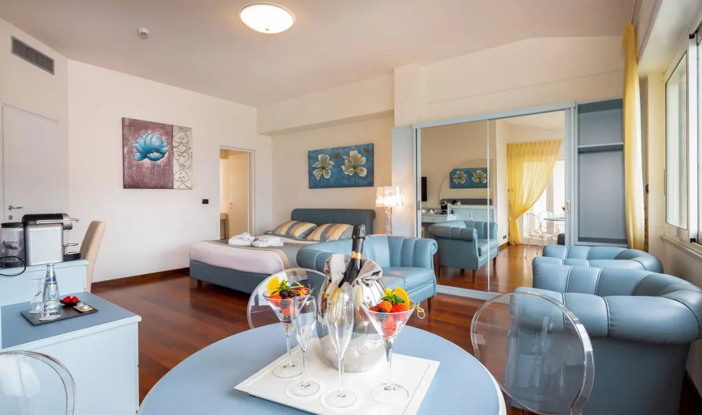 Nell'immagine, l'interno di una suite con pavimeto in legno, 3 poltrone in pelle azzurra, in primo piano un tavolino con delle coppe di frutta fresca e una bottiglia di champagne e, sullo sfondo, un letto matrimoniale