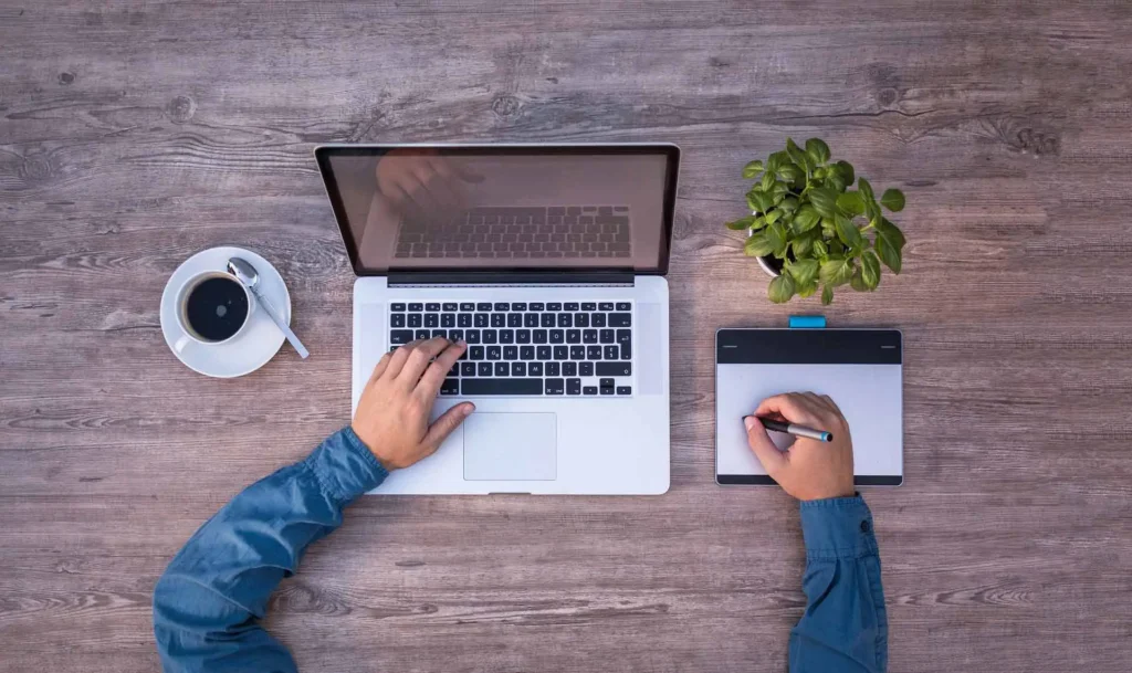 Nell'immagine un tavolo sui cui sono appoggiati un laptop, una tazza di caffè, un tablet e una piantina. Si vedono due mani che lavorano sul laptop