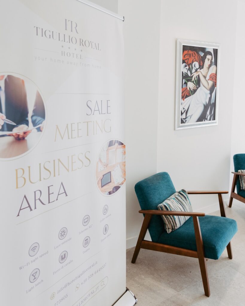 Roll-up promozionale del Tigullio Royal Hotel dedicato a “Sale Meeting Business Area”, posizionato accanto a due poltrone imbottite color verde petrolio con braccioli in legno; parete chiara con quadro decorativo raffigurante una figura stilizzata e fiori rossi, ambiente luminoso e moderno.